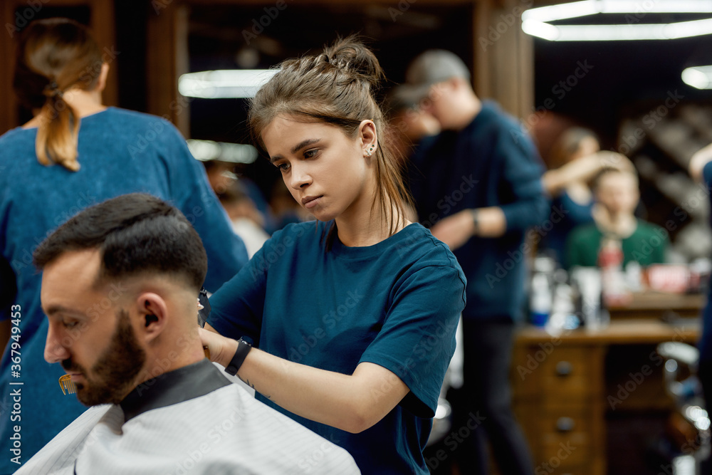 Obraz premium Focused professional barber girl making trendy haircut for a young bearded man sitting in barber shop chair. Side view. Focus on a girl