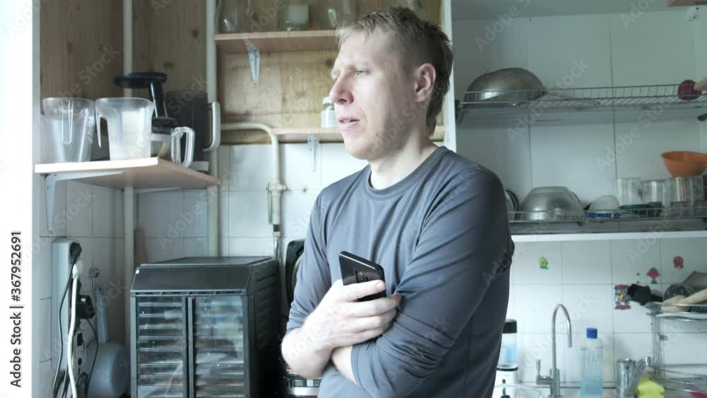 young thoughtful caucasian man in grey turtleneck standing at old soviet kitchen, speaking to himself, holding smartphone in hands. shelfs with dishes in background young thoughtful caucasian man in grey turtleneck standing at old soviet kitchen, speaking to himself, holding smartphone in hands. shelfs with dishes in background