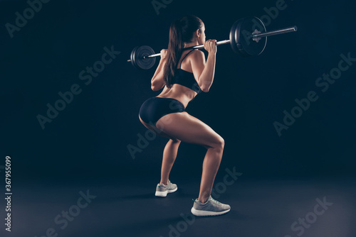 Full body side behind photo active short sport suit lady shoulders heavy barbell fitness doing squats isolated black background