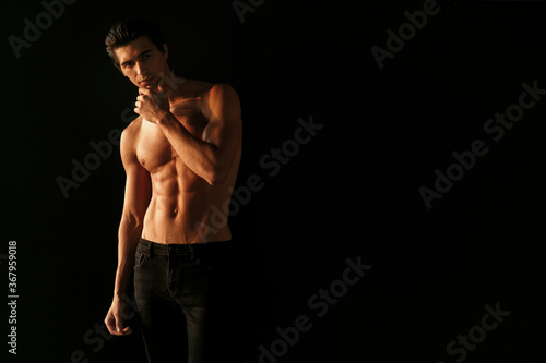 Strong athletic handsome young man with naked torso, showing his six abs on dark black background.