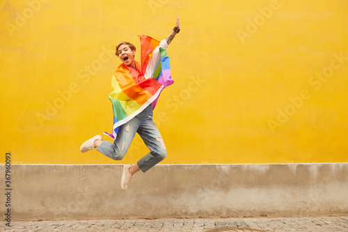 Portrait of young happy lesbian with colored flag on her shoulders jumping outdoors