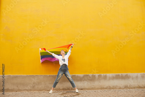 Young happy lesbian holding colored flag and smiling against the yellow wall in the city outdoors