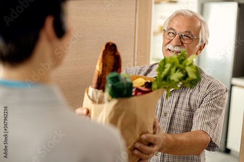 Tableau sur toile Happy senior man taking bag with groceries from a courier at front door