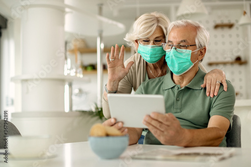 Mature couple with face masks making video call over touchpad at home.