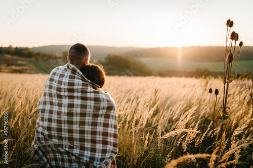 Young couple hugging, standing back, people covered with blanket, at sunset in autumn an outdoor. at field grass on background of sun. Concept of friendly family. full length. Close Up.