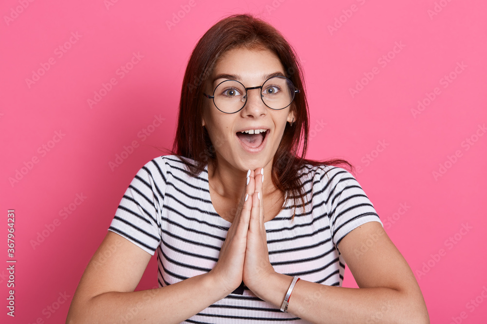 Indoor shot of beautiful young female with positive expression, keeps palms together, has astonished expression, poses against rosy background, keeping mouth widely opened. Indoor shot of beautiful young female with positive expression, keeps palms together, has astonished expression, poses against rosy background, keeping mouth widely opened.