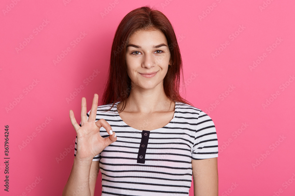 Portrait of happy young brunette girl showing ok gesture with fingers, looking at camera with charming smile, has excellent news, wearing striped t shirt. Portrait of happy young brunette girl showing ok gesture with fingers, looking at camera with charming smile, has excellent news, wearing striped t shirt.