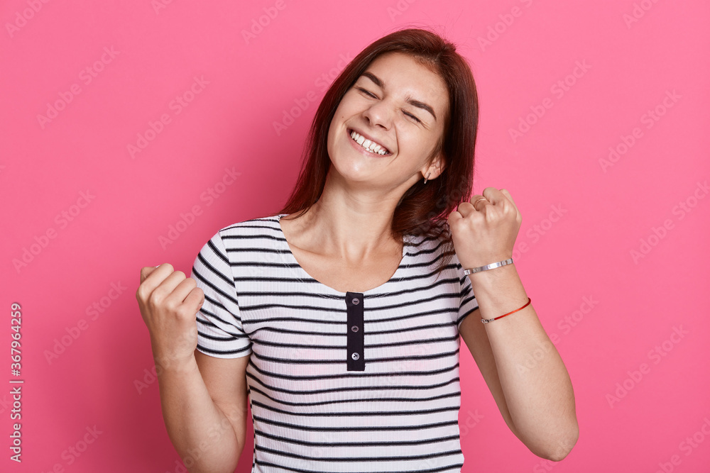 Excited cheerful female with joyful expression, cheers and clenches fists, celebrating her success, poses against pink background, dresses casual white and black striped t shirt. Excited cheerful female with joyful expression, cheers and clenches fists, celebrating her success, poses against pink background, dresses casual white and black striped t shirt.