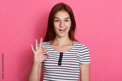 Young beautiful woman doing ok sign with fingers and showing her tounge  looking directly at camera with funny expression  posing isolated over pink background.