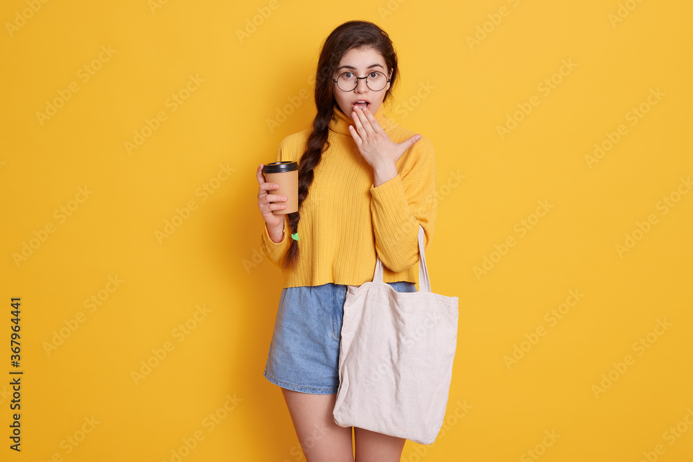 Female holding reusable coffee mug and cotton bag while posing isolated over yellow background, having astonished facial expression, covering her widely opened mouth with palm. Female holding reusable coffee mug and cotton bag while posing isolated over yellow background, having astonished facial expression, covering her widely opened mouth with palm.