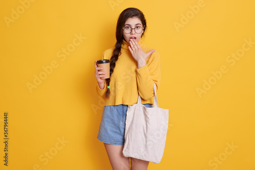 Female holding reusable coffee mug and cotton bag while posing isolated over yellow background, having astonished facial expression, covering her widely opened mouth with palm.