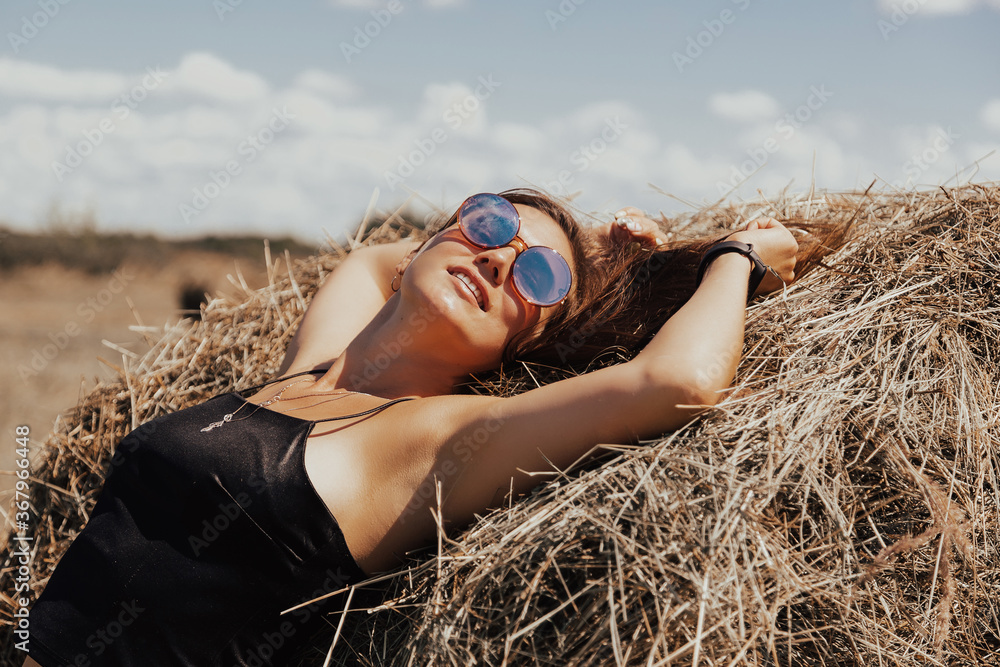 Beautiful young girl with long hair in sunnglasses posing on a wheat field near hay bales.