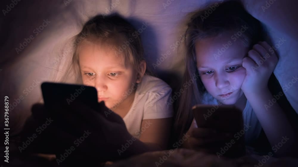 Two little girls with smatfonds do not sleep under a blanket. Children's insomnia and Internet addiction to devices and gadgets Two little girls with smatfonds do not sleep under a blanket. Children's insomnia and Internet addiction to devices and gadgets
