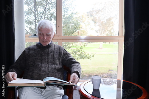 Senior male reading a book as one of his past time while while isolating. Behind him is a big window that shows a big yard.