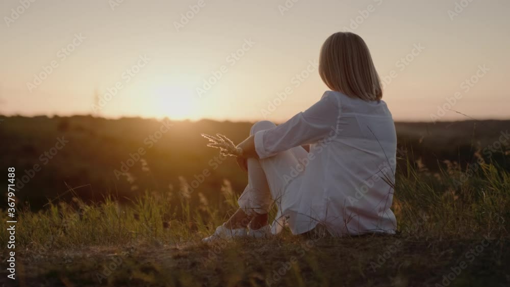 A woman in a white suit sits on a hill, admiring the sunset