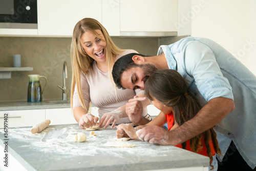 Happy excited mom and dad having fun while teaching daughter to make dough at kitchen table. Young couple and their girl baking buns or pies together. Family cooking concept