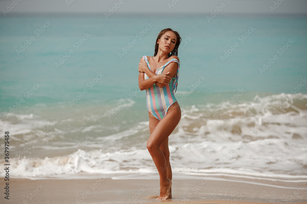 Fototapeta premium Beautiful and young girl in a swimsuit posing on the ocean and sea with blue waves. Woman in a swimsuit model and fashion posing