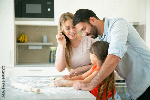 Mom and dad teaching kid to knead dough on kitchen table with flour messy. Young couple and their girl baking buns or pies together. Family cooking concept