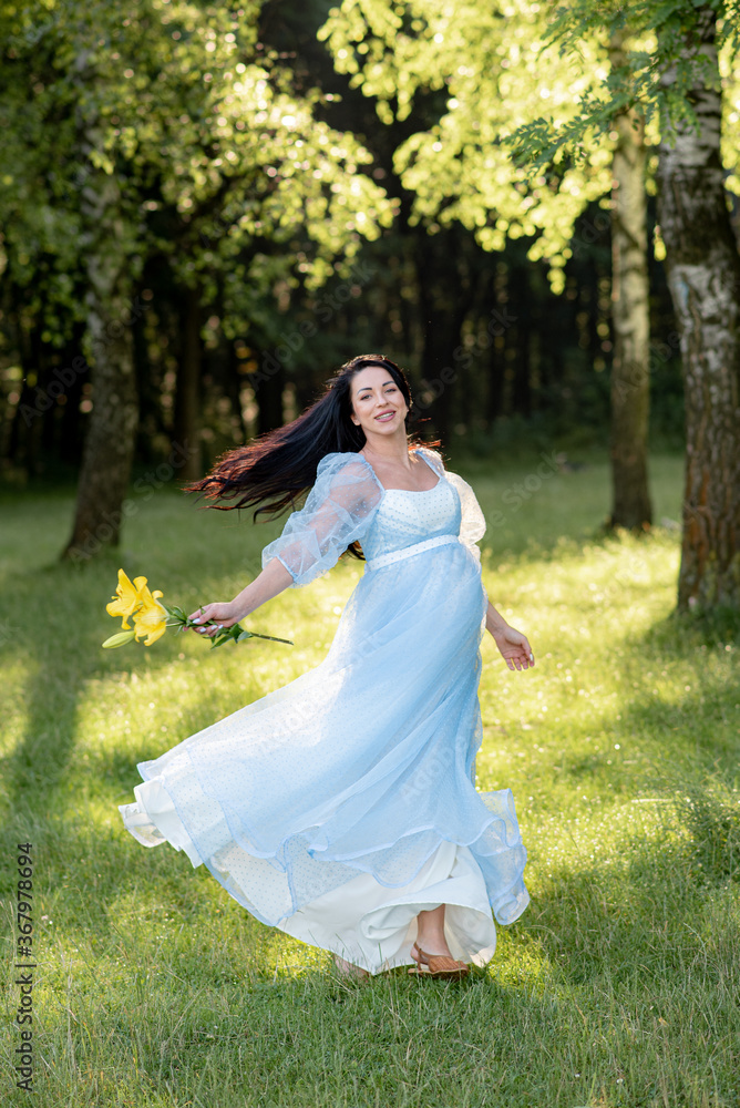 Pregnant woman posing in a blue dress on a background of green trees.