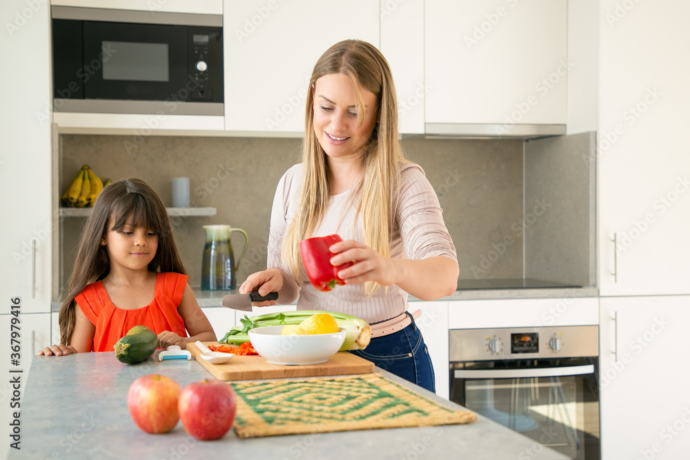 Mom showing daughter how to cook salad for dinner. Girl and her mother cutting vegs on kitchen counter. Medium shot, copy space. Family cooking concept