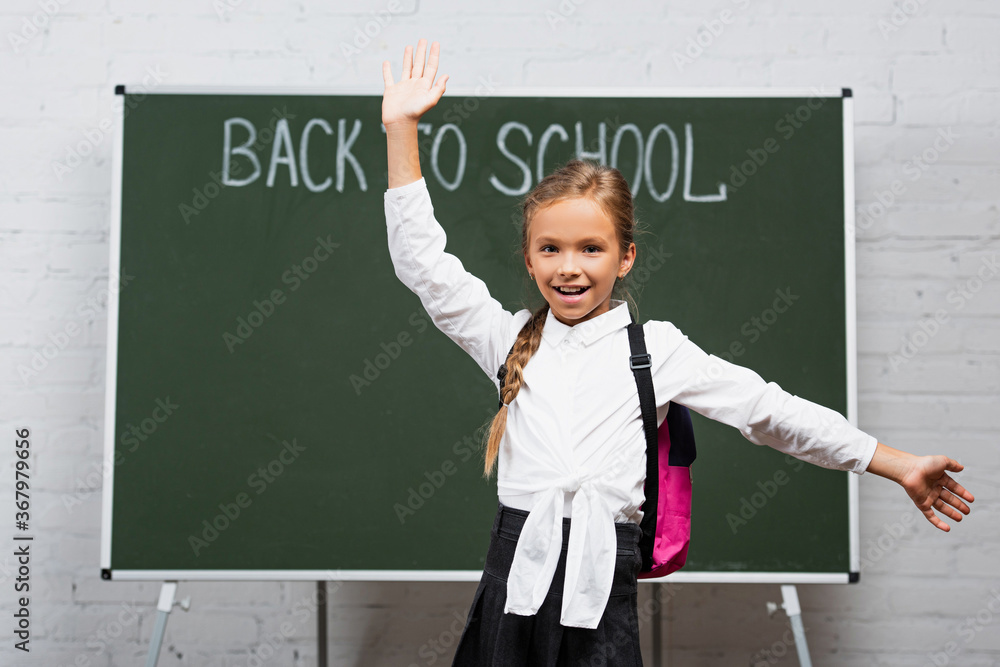 selective focus of happy schoolgirl with backpack and open arms smiling at camera near chalkboard with back to school lettering