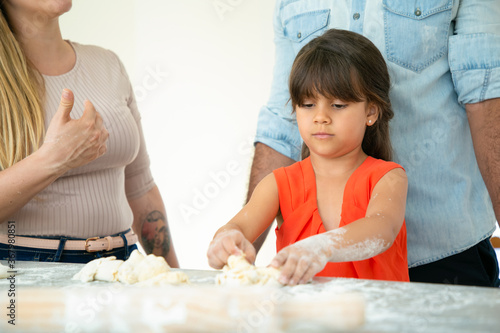 Mom and dad watching daughter making dough on kitchen table with flour messy. Young couple and their girl baking buns or pies together. Family cooking concept