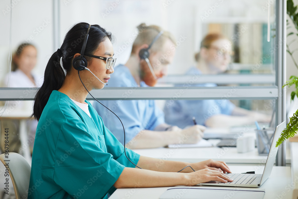 Asian young operator in uniform and headset sitting at the table and typing on laptop at hospital Asian young operator in uniform and headset sitting at the table and typing on laptop at hospital