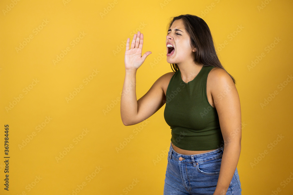 Young beautiful woman standing over yellow isolated screaming with her hand in the air Young beautiful woman standing over yellow isolated screaming with her hand in the air