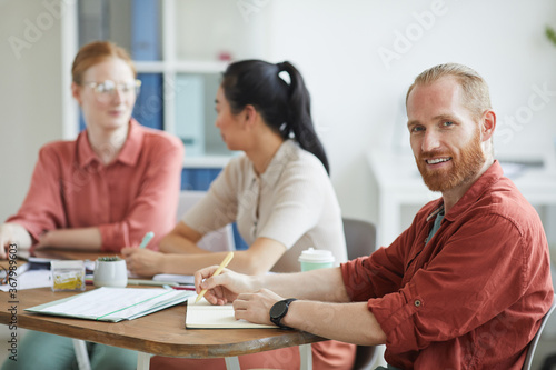 Portrait of young bearded businessman looking at camera while sitting at the table with his colleagues during business meeting