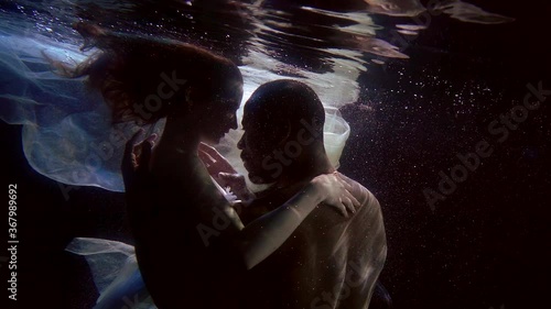 man and woman are having sex underwater in dark depth of pool, brawny man is embracing girlfriend photo