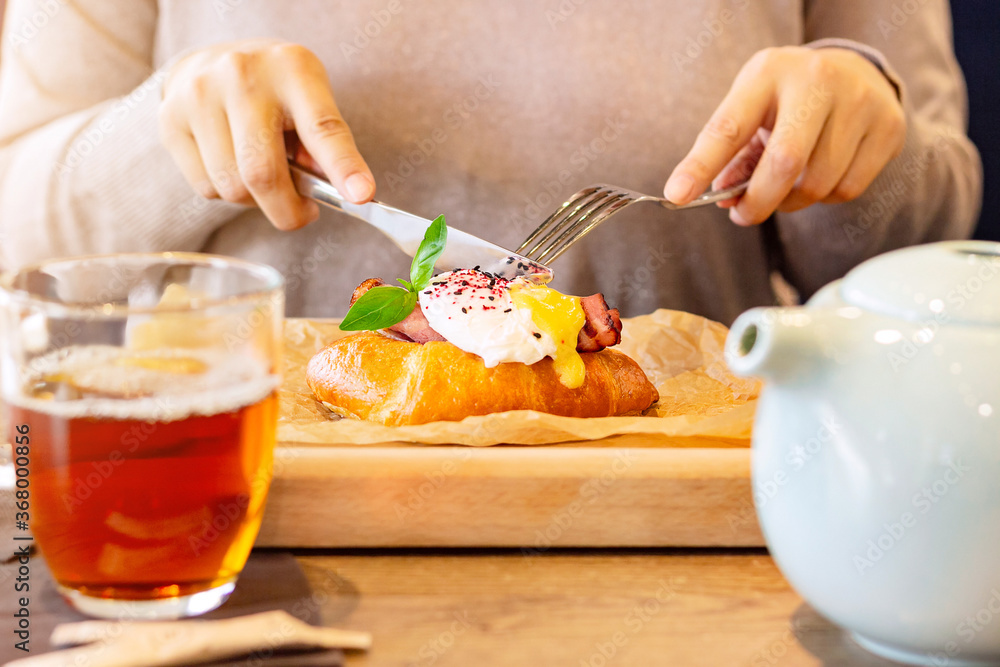 Woman has breakfast in a restaurant, egg benedict with croissant on a wooden plate under baking paper, brunch food style Woman has breakfast in a restaurant, egg benedict with croissant on a wooden plate under baking paper, brunch food style