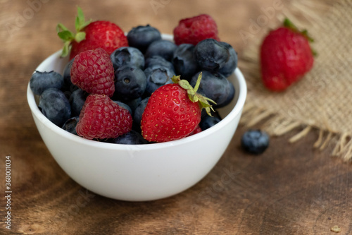 Set of summer berries in a small white bowl on a napkin and wooden background. Blueberries  raspberries  and strawberries are a varied healthy snack. Yummy.
