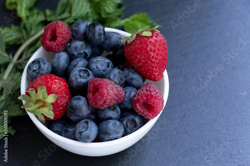 Set of summer berries in a small white bowl on a blackboard. Blueberries  raspberries  and strawberries are a varied healthy snack. Yummy.