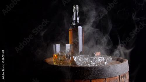 A glass of whiskey with ice cubes, a bottle and a cigar in the smoke on a wooden barrel