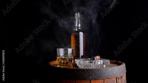 A glass of whiskey with ice cubes, a bottle and a cigar in the smoke on a wooden barrel
