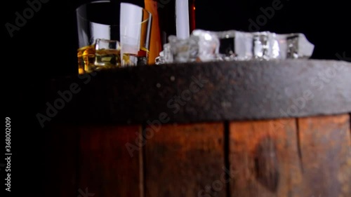 A glass of whiskey with ice cubes, a bottle and a cigar in the smoke on a wooden barrel