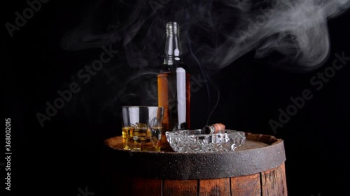 A glass of whiskey with ice cubes, a bottle and a cigar in the smoke on a wooden barrel