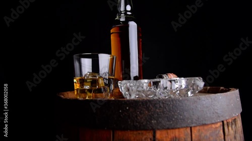 A glass of whiskey with ice cubes, a bottle and a cigar in the smoke on a wooden barrel