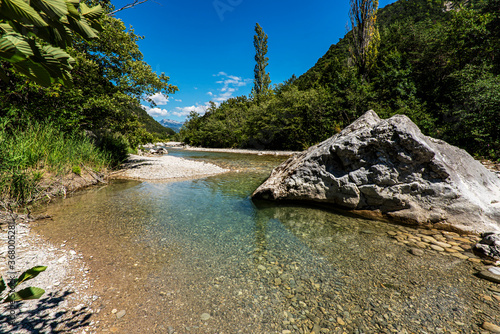 Turquoise water of the french Drome river running between pebbly river banks and green forest with high mountains on the background.