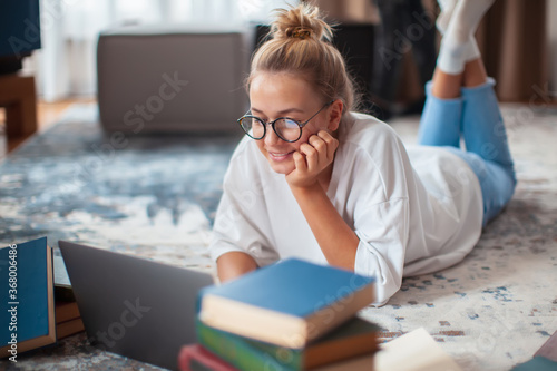 Obraz na plátně Cute teen girl student in casual clothes, glasses lying on floor at laptop with books