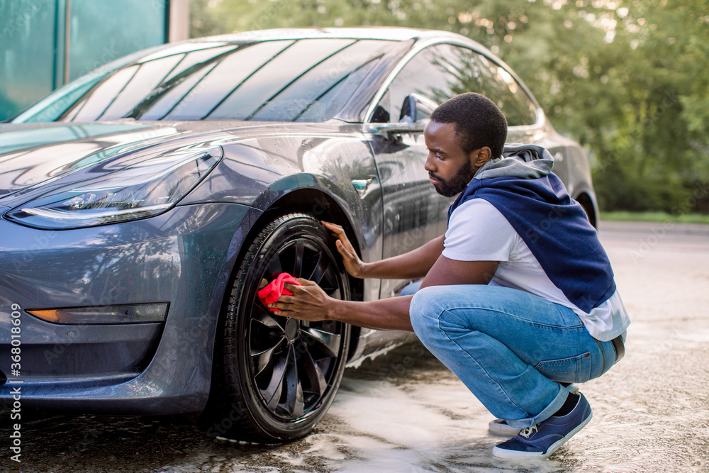 Car wash service outdoors. Car wash self-service concept. Handsome African guy wiping wheel of his modern luxury electric car with red cloth during the washing process outdoors Car wash service outdoors. Car wash self-service concept. Handsome African guy wiping wheel of his modern luxury electric car with red cloth during the washing process outdoors