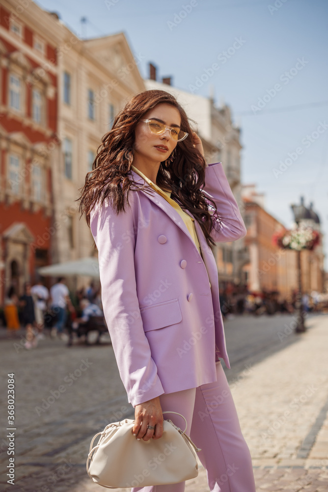 Outdoor fashion portrait of elegant woman wearing lilac suit, holding trendy big white leather pouch handbag, posing in street of European city