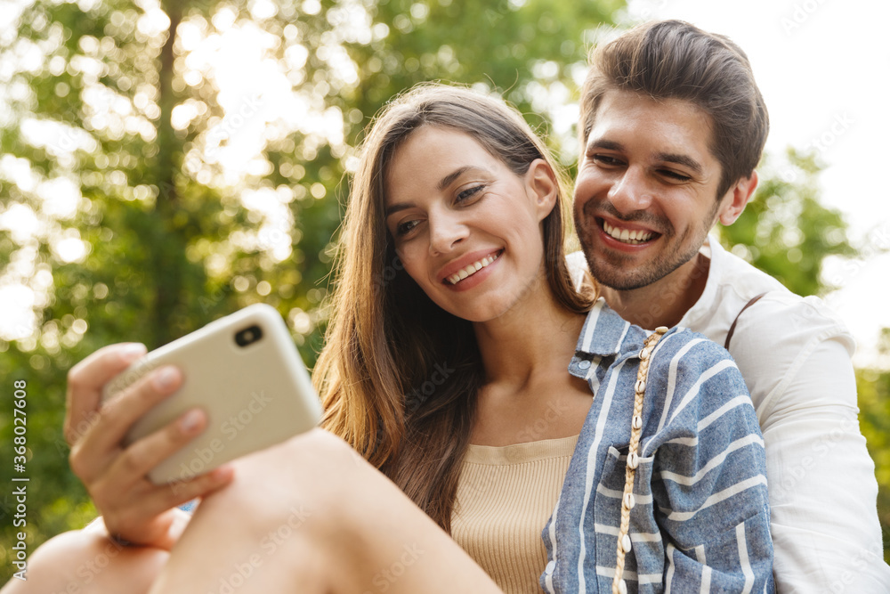 Image of young happy couple smiling and holding cellphone in park Image of young happy couple smiling and holding cellphone in park