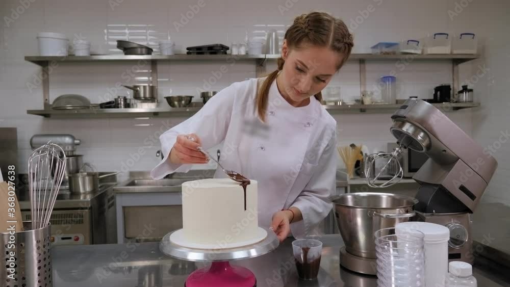 Close-up of a pastry chef pouring liquid chocolate from a spoon on a white cream sponge cake. Decorating a cake with cream and chocolate in a professional pastry kitchen. Close-up of a pastry chef pouring liquid chocolate from a spoon on a white cream sponge cake. Decorating a cake with cream and chocolate in a professional pastry kitchen.