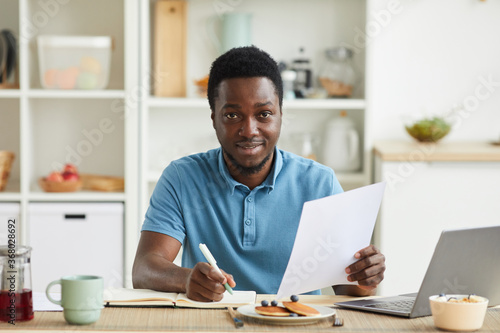Portrait of African young man planning his day and making notes in note pad while sitting at the table in the kitchen