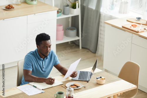 African young man sitting at the table in front of laptop and working with documents in the kitchen © AnnaStills