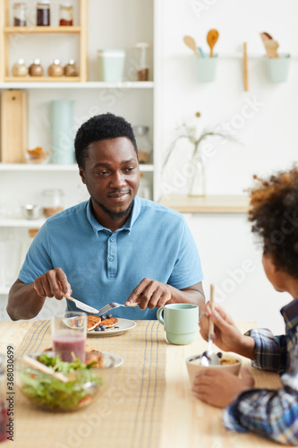 African young father has breakfast together with his son at the table in the kitchen they smiling and talking