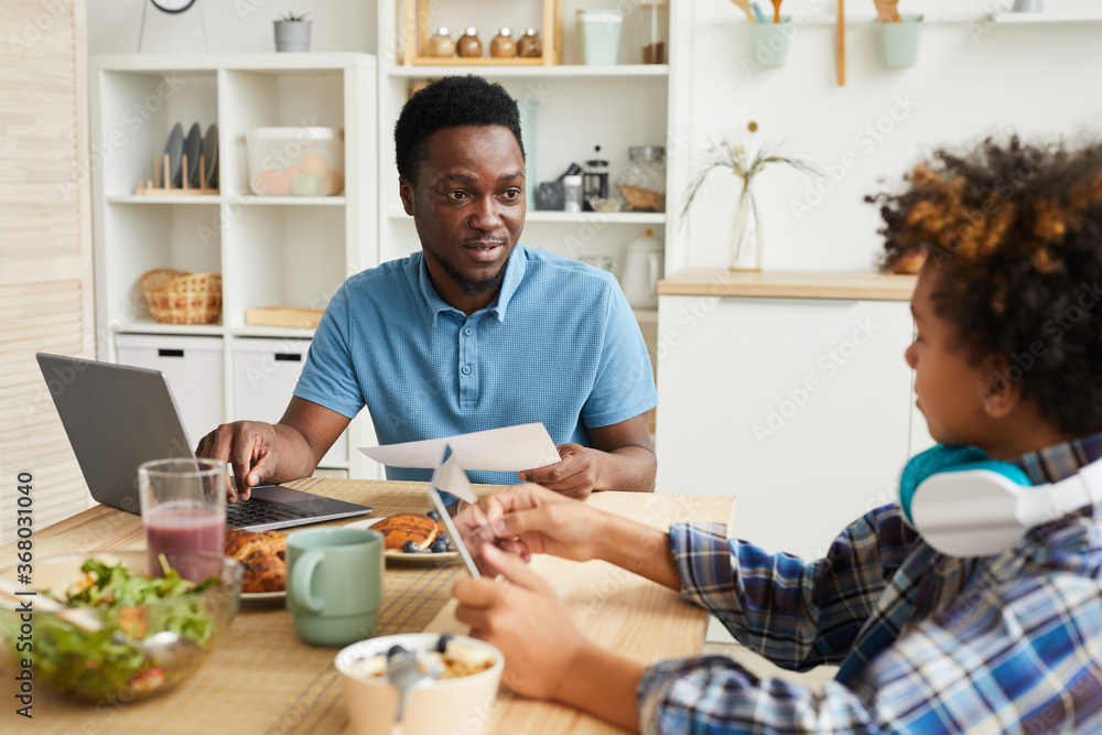Φωτογραφία African young father sitting at the table in front of laptop and talking to his