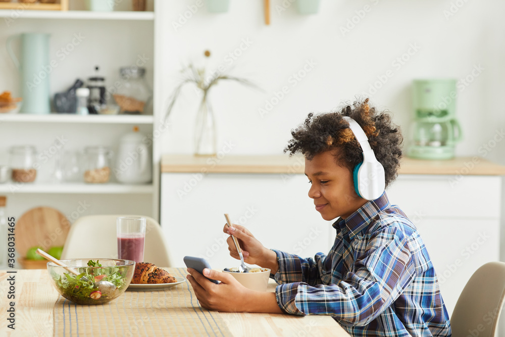 African boy in headphones using his mobile phone while eating breakfast in the kitchen African boy in headphones using his mobile phone while eating breakfast in the kitchen