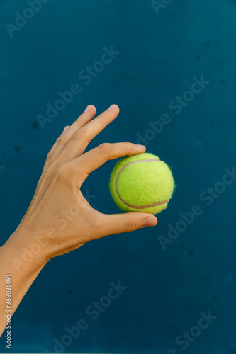  Yellow tennis ball in hand on the background of blue wall. Doing ok sign with hand and fingers. Successful expression.